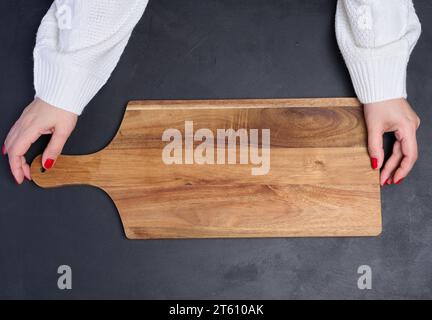 Planche à découper en bois vide dans les mains des femmes sur une table en bois noir, vue de dessus Banque D'Images