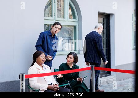 Foule de personnes diverses assis à l'extérieur à la porte du centre commercial qui attendent dans la file pour magasiner pour les ventes saisonnières des fêtes. Les clients anxieux à l'entrée du magasin de détail attendent les offres du Black Friday Banque D'Images