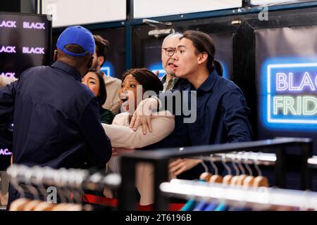 Foule de personnes diverses debout à l'entrée du magasin de vêtements devenant folles pendant le Black Friday. Les acheteurs se disputent les combats avec l'homme afro-américain gardien de sécurité en attendant dans la queue. Chasse aux bonnes affaires Banque D'Images