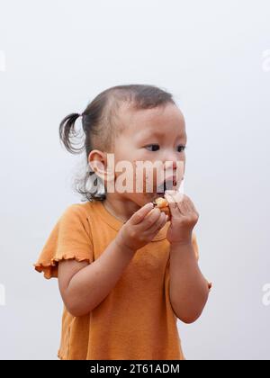 Mignonne petite fille asiatique mangeant un beignet sucré ou du pain au chocolat, la bouche recouverte de chocolat. Banque D'Images