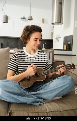 Portrait vertical d'une jeune femme heureuse jouant ukulélé, apprend un nouvel instrument de musique, s'assoit sur un canapé à la maison, souriant de joie Banque D'Images