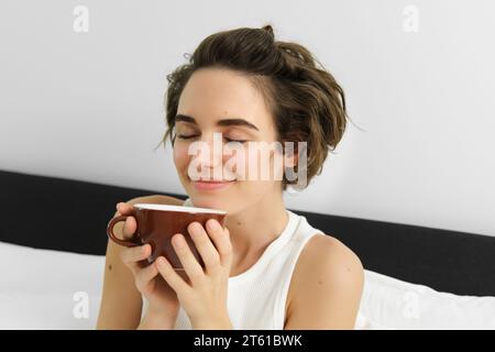 Gros plan portrait de femme sentant son café, prenant la tasse du matin de boisson aromatique préférée, ferme les yeux et souriant, assis sur le lit Banque D'Images