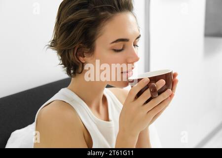 Portrait de jeune femme se réveillant, buvant du café au lit, sentant l'arôme préféré Banque D'Images