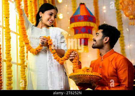 Large plan de jeune homme indien aidant sa femme pour décorer des fleurs pendant la célébration du festival Diwali en se parlant à la maison - concept de Banque D'Images