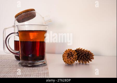 Thé noir dans une tasse en verre et théière en verre fraîchement infusée sur une serviette et des pommes de pin avec fond blanc Banque D'Images