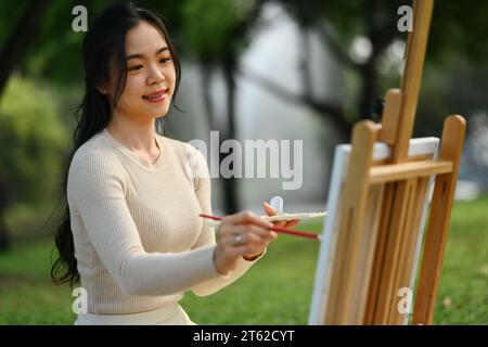 Jeune femme souriante peignant un tableau dans le parc. Concept de pleine conscience, d'art thérapie et de loisirs créatifs Banque D'Images