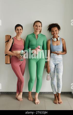 Portrait vertical pleine longueur de trois femmes diverses portant des vêtements d'entraînement et souriant à la caméra dans la salle de gym Banque D'Images