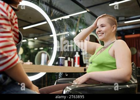 Portrait de jeune femme confiante avec buzzcut souriant joyeusement dans le salon de beauté et bavardant avec le coiffeur Banque D'Images
