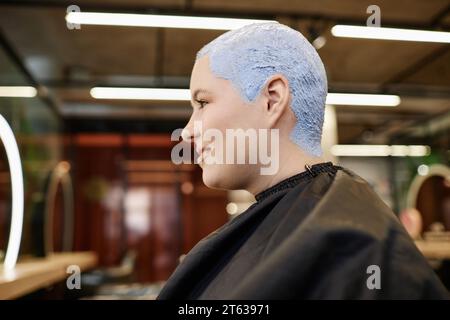 Portrait de vue latérale de jeune femme souriante avec les cheveux buzzcut blanchis regardant dans le miroir dans le salon de beauté Banque D'Images