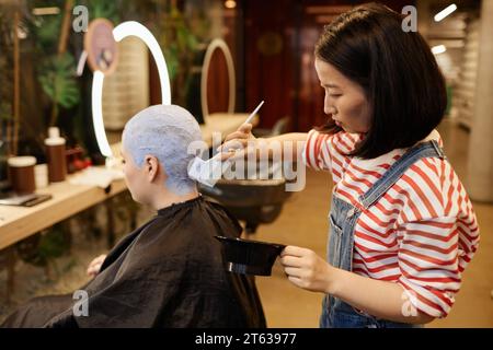 Portrait de vue latérale de jeune femme asiatique comme coiffeur appliquant de l'eau de Javel aux cheveux courts buzzcut dans le salon de beauté Banque D'Images