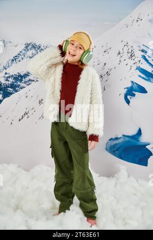 petite fille en tenue d'hiver élégante avec des écouteurs écoutant de la musique et souriant joyeusement, la mode Banque D'Images