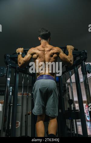 Vue arrière d'un homme musclé faisant des tractions sur un bar dans une salle de gym faiblement éclairée mettant en valeur ses muscles du dos bien définis Banque D'Images