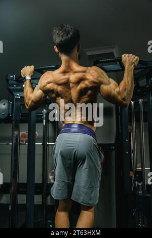 Vue arrière d'un homme musclé faisant des tractions sur un bar dans une salle de gym faiblement éclairée mettant en valeur ses muscles du dos bien définis Banque D'Images