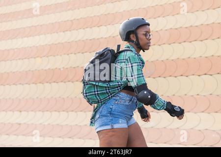 Vue latérale de la jeune femme afro-américaine dans l'équipement de protection portant sac à dos tout en patinant à roulettes au skate Park Banque D'Images