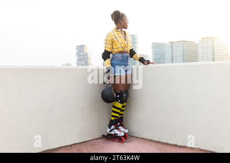 Pensive jeune femme afro-américaine en patins à roulettes tenant le casque tout en se tenant au skate Park contre le ciel clair Banque D'Images