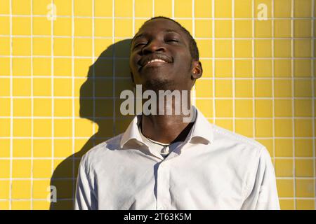 Joyeux jeune homme noir riant contre un mur carrelé jaune vibrant, avec le soleil projetant une ombre à côté de lui Banque D'Images