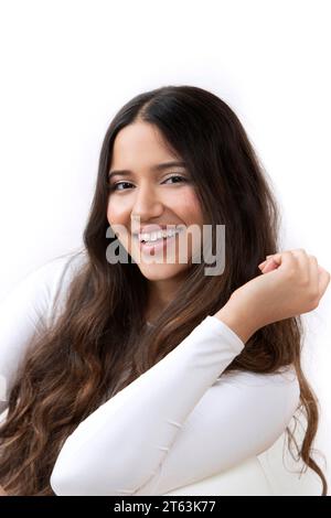 Portrait rapproché d'une jeune femme souriante avec de longs cheveux foncés ondulés portant un haut blanc sur un fond blanc Banque D'Images