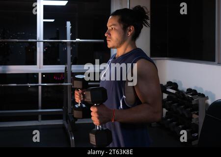 Vue latérale d'un homme concentré soulève des haltères dans une salle de sport faiblement éclairée avec les lumières nocturnes de la ville scintillant à travers la fenêtre derrière lui Banque D'Images