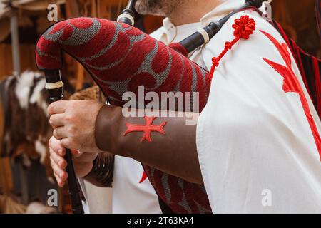 Gros plan d'un bras de reconstituteur médiéval anonyme drapé dans un tissu à motifs rouges tenant un instrument de musique avec un emblème de croix rouge tatoué sur le FO Banque D'Images