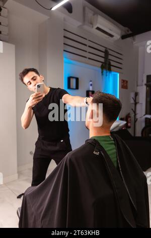 Barber prendre une photo avec un smartphone tout en ajustant les cheveux d'un client masculin, mettant en valeur l'environnement moderne et bien éclairé du salon. Banque D'Images