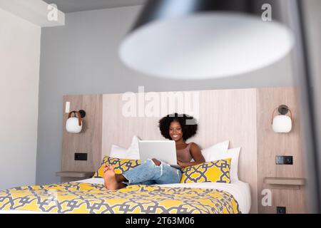 Joyeuse femme noire avec des salons de cheveux bouclés sur un lit moderne noyé dans son ordinateur portable entouré par la décoration contemporaine de la chambre Banque D'Images