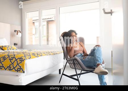 Femme noire détendue avec les cheveux bouclés assis sur une chaise moderne par la fenêtre parlant au téléphone dans une chambre bien éclairée avec un décor contemporain Banque D'Images