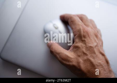 Gros plan d'une main âgée d'une personne anonyme saisissant une souris blanche d'ordinateur sur une surface lisse floue Banque D'Images