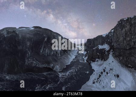 Vue panoramique d'une rivière gelée coulant à travers un canyon enneigé avec des falaises abruptes, le tout sous un ciel étoilé sombre en Islande Banque D'Images