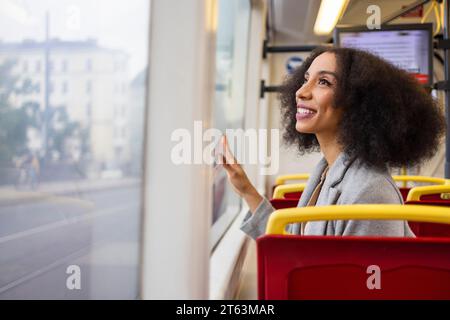 Joyeuse femme ethnique avec les cheveux bouclés regarde par la fenêtre d'un bus de la ville tout en admirant la vue extérieure dans la rue Banque D'Images