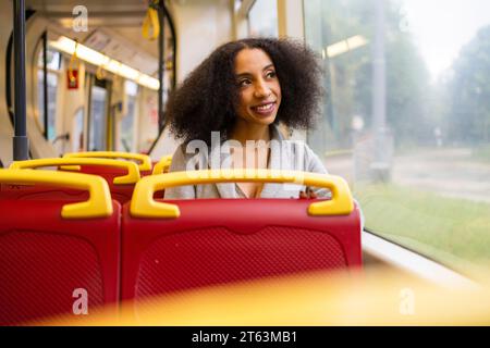 Joyeuse femme ethnique avec les cheveux bouclés regarde par la fenêtre d'un bus de la ville tout en admirant la vue extérieure dans la rue Banque D'Images