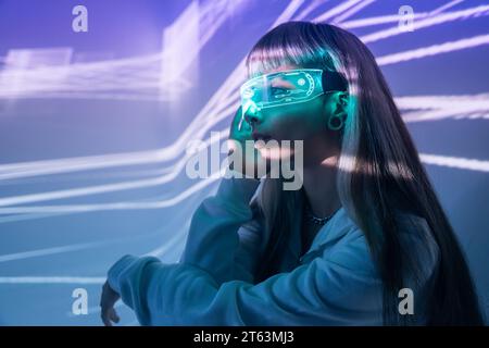 Vue latérale de la jeune femme hypnotisée par des lunettes LED VR modernes et immergée dans l'expérience de réalité virtuelle sur des lumières de projecteur éclairées Banque D'Images