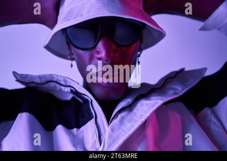 Gros plan de beau jeune homme afro-américain confiant avec piercing sur le nez et boucles d'oreilles portant chapeau et lunettes de soleil debout sur fond violet W Banque D'Images