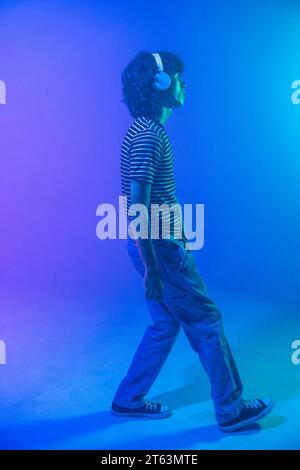 Un jeune homme aux cheveux bouclés et à la chemise rayée, portant des écouteurs, marche dans une ambiance bleue dégradée, perdu dans son univers musical Banque D'Images