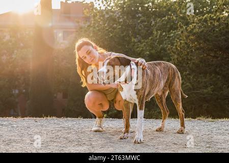 Une jeune femme s'accroupe dans un parc, souriant alors qu'elle embrasse un chien américain Stanford brun et blanc baigné dans la lumière chaude du coucher du soleil, avec des arbres dans le dos Banque D'Images