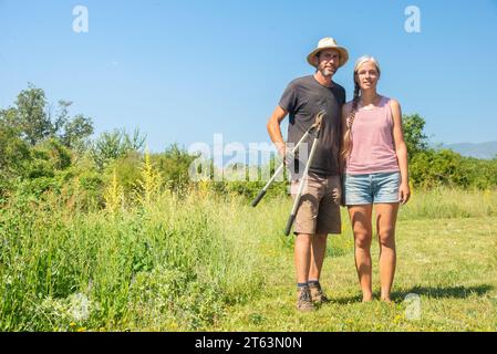Corps entier d'un homme dans un chapeau de paille debout à côté d'une femme dans un débardeur rayé, tenant des outils de jardinage tout en regardant la caméra, avec un champ vibrant Banque D'Images