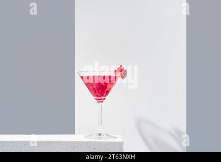 Verre rempli de cocktail de grenade rouge servi avec des graines de grenade sur fond gris et blanc, projetant de l'ombre Banque D'Images