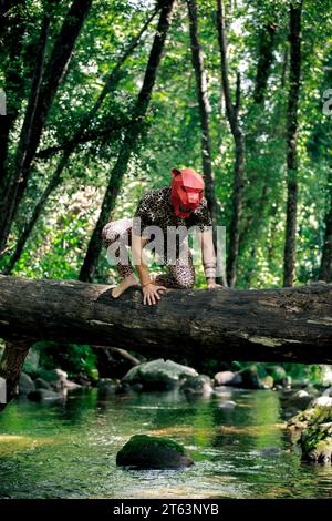 Mâle anonyme habillé comme guépard avec masque rouge marchant à quatre pattes sur le tronc d'arbre contre les arbres verts dans les gorges dans la nature Banque D'Images