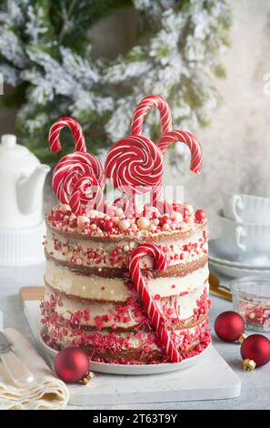 Gâteau de pain d'épice de Noël en couches décoré de Lolli POPS rouges, confettis colorés sur un support de gâteau blanc, boules de Noël, branches de sapin sur béton blanc Banque D'Images