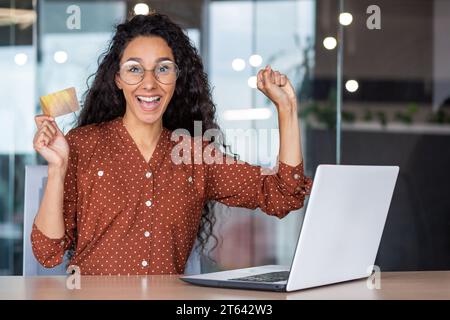 Femme attractive hispanique est ravie de l'achat qu'elle a fait des achats en ligne. Curly brunette dans les verres s'assoit à la table devant l'ordinateur portable, tient la carte dans ses mains et fait joyeusement le paiement en ligne. Banque D'Images