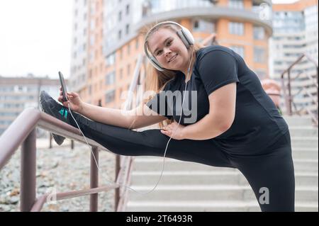 Grosse jeune femme faisant des exercices d'étirement et écoutant de la musique avec des écouteurs à l'extérieur Banque D'Images