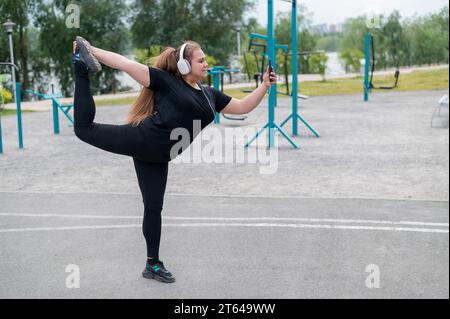 Belle grosse jeune femme dans les écouteurs fait un patineur artistique et prend un selfie. Une jeune fille souriante écoute de la musique et se réchauffe avant de faire du jogging Banque D'Images