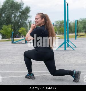 Une belle grosse fille dans un survêtement noir est engagée dans la remise en forme sur le terrain de sport. Une jeune femme se jette à l'extérieur par une chaude journée d'été. Sain Banque D'Images