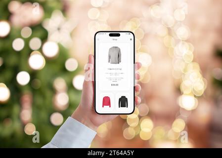 La main de la femme tient un téléphone mobile avec une application e-commerce moderne, engagée dans les achats de Noël. Le bokeh festif de lumières de sapin de Noël crée une holida Banque D'Images