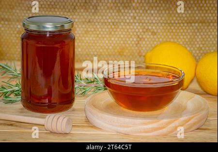 Pot de miel, distributeur, un bol avec du miel sur une assiette en bois, une branche de romarin et deux citrons sur bois avec un vrai nid d'abeille en arrière-plan Banque D'Images