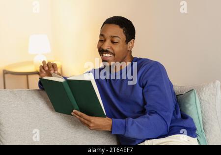 Homme millénaire noir lit livre de poche assis dans la maison contemporaine Banque D'Images