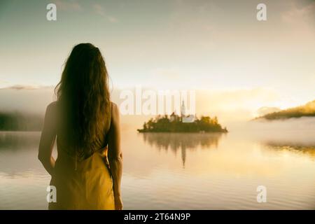 Femme en robe jaune regardant un lever de soleil parmi le brouillard sur les rives du lac de Bled, Slovénie VII Banque D'Images
