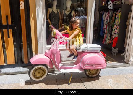 Jeune fille assise sur un scooter rose vespa devant une boutique de vêtements, Calle Don Gerónimo, Ávila, Castille-et-León, Royaume d'Espagne Banque D'Images