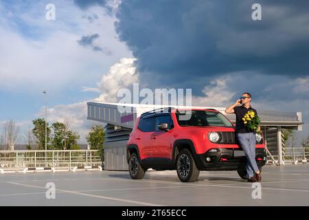 Un homme avec un bouquet de fleurs parle au téléphone tout en se tenant près de la voiture dans le parking. Concept de voyage Banque D'Images