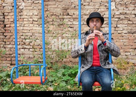 Un musicien de rue sur une balançoire pour enfants joue d'un instrument de musique en bois Banque D'Images