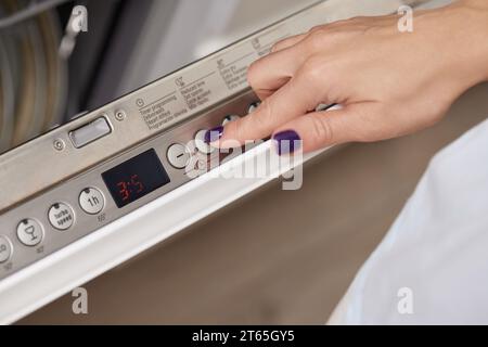 main féminine tournant sur la machine lave-vaisselle dans la cuisine, femme choisissant le programme de mode sur le panneau de commande du lave-vaisselle. Banque D'Images